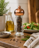A Bisetti Clear copper pepper mill grinder with a clear glass body filled with whole peppercorns. Bisetti-USA