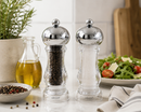 Bisetti capri pepper and salt mills with shiny chrome tops, shown on a kitchen counter with a salad, olive oil, and peppercorns in a real home environment.