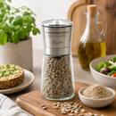 Bisetti sunflower seeds grinder filled with seeds, styled on a wooden board with basil, olive oil, and bowls of food in a kitchen setting.
