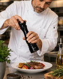 A chef in a white uniform grinds black pepper with Bisetti Sorrento mill over a plated steak with roasted vegetables in a kitchen setting.